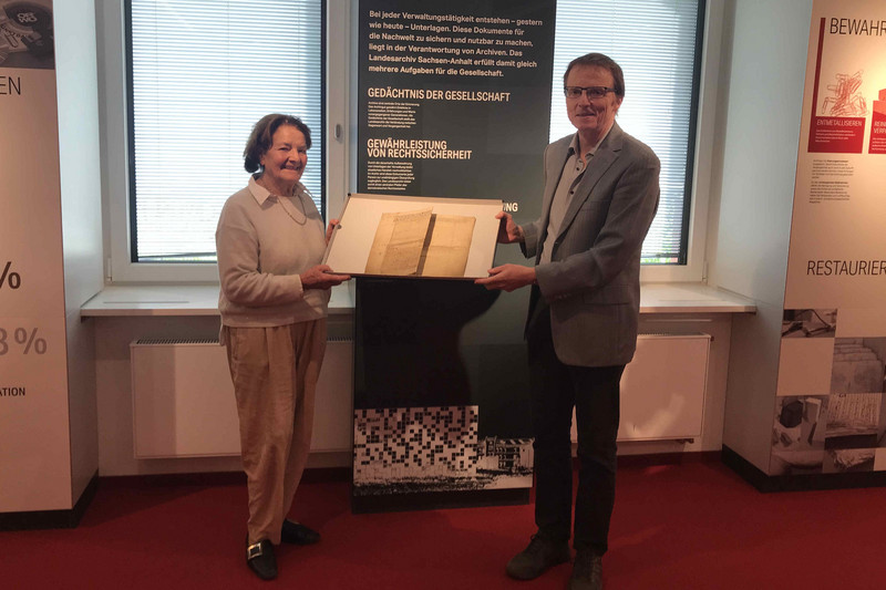 Eine Frau und ein Mann vor beschrifteten Rollups einer Ausstellung im Landesarchiv Sachsen-Anhalt, in den Händen ein mittelalterliches Nekrologblatt haltend.