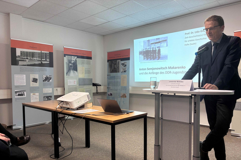 Man vor Rednerpult, im Hintergrund die Vortragspräsentation und Ausstellungsrollups